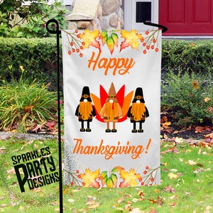 Op de afbeelding: Een witte tuin vlag met een herfstthema. Het toont drie kabouters die voor een kalkoen staan met de tekst "Happy Thanksgiving!" in oranje letters. De vlag is versierd met herfstbladeren en bessen.