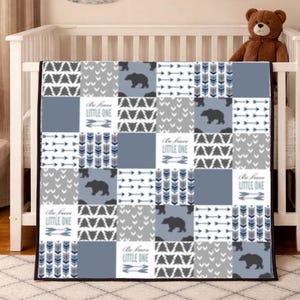May include: A patchwork baby blanket with a bear and arrow pattern in shades of grey, blue, and white. The blanket features the text "Be brave LITTLE ONE". The blanket is displayed on a white cot.