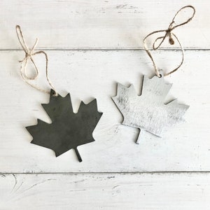 May include: Two metal maple leaf ornaments with twine hangers. One leaf is painted silver and the other is black.