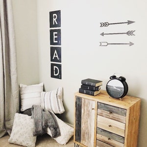 May include: A wooden dresser with four drawers and a cabinet door. The dresser has a rustic, reclaimed wood look and is painted in shades of brown, gray, and white. The dresser has black metal hairpin legs. The dresser is decorated with a stack of books and a black alarm clock. The wall behind the dresser has a black metal sign that reads "READ" and three black metal arrows.