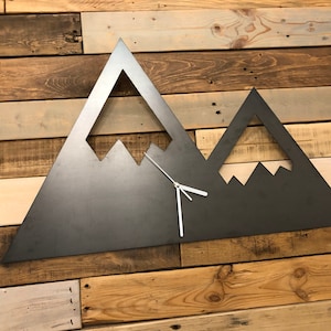 Peut inclure: Une horloge murale en métal en forme de chaîne de montagnes avec des aiguilles blanches. L'horloge est montée sur un mur en panneaux de bois.