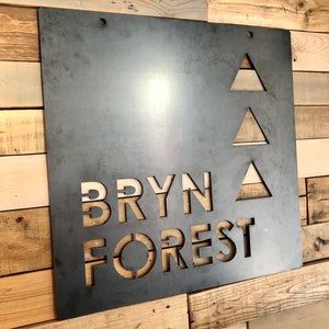 May include: A metal sign with the words "BRYN FOREST" cut out in a stylized font. Three triangles are cut out above the text.