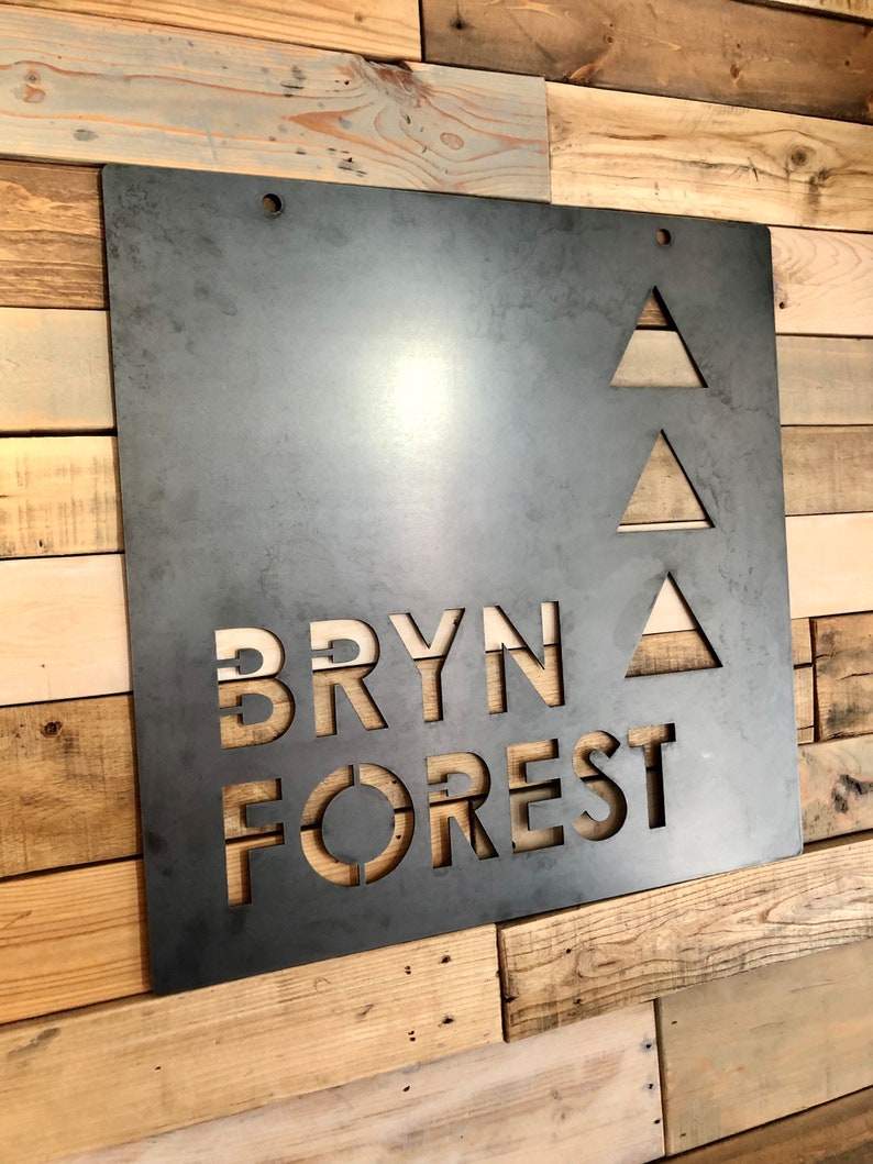 May include: A metal sign with the words "BRYN FOREST" cut out in a stylized font. Three triangles are cut out above the text.