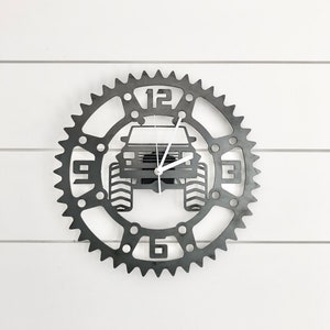 May include: A metal clock with a gear design featuring a silhouette of a jeep in the centre. The clock face has black numbers and white hands.