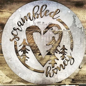 May include: A round metal sign with the words "scrambled" and "home" written in a cursive font. The sign features a heart shape with a tree design inside.