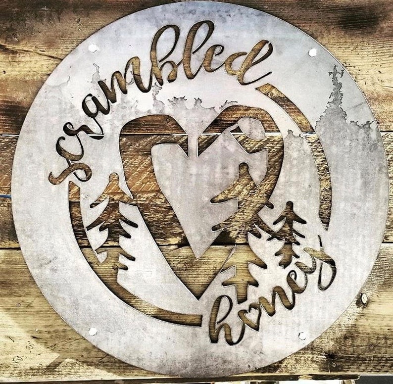 May include: A round metal sign with the words "scrambled" and "home" written in a cursive font. The sign features a heart shape with a tree design inside.