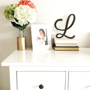 May include: A white dresser with two drawers and a gold vase with pink and green flowers. A white picture frame with a photo of a woman holding a baby is on the dresser. A black wooden letter L is on top of a stack of books.