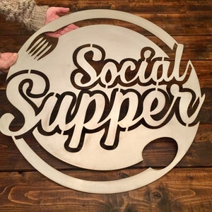 May include: A round metal sign with the words "Social Supper" cut out in a stylized font. A fork and knife are also cut out in the design.