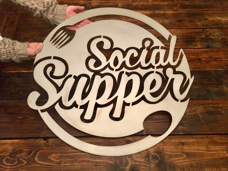 May include: A round metal sign with the words "Social Supper" cut out in a stylized font. A fork and knife are also cut out in the design.