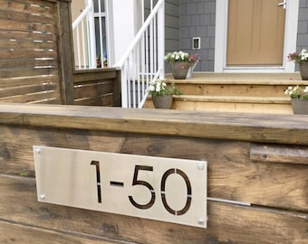 May include: A stainless steel house number plaque with the number "1-50" cut out. The plaque is mounted on a wooden fence.