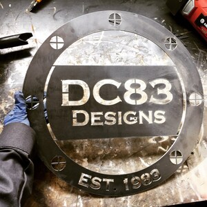 May include: A black metal round sign with the text "DC83 Designs Est. 1983" in white lettering. The sign has a distressed look and is surrounded by a decorative border.