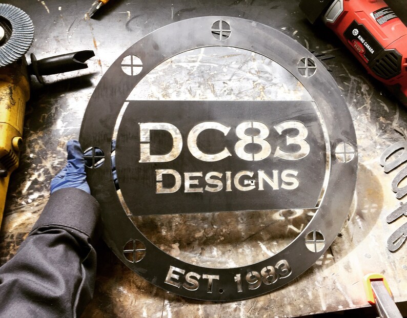May include: A black metal round sign with the text "DC83 Designs Est. 1983" in white lettering. The sign has a distressed look and is surrounded by a decorative border.