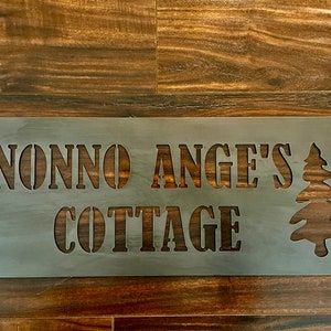 May include: A metal sign with the text "Nonno Ange's Cottage" and a stylized pine tree design.