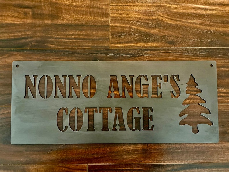 May include: A metal sign with the text "Nonno Ange's Cottage" and a stylized pine tree design.