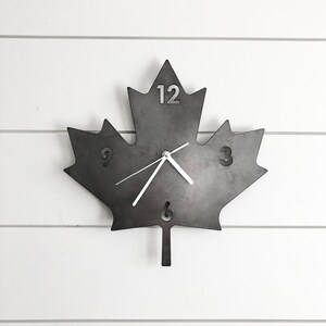 Peut inclure: Une horloge en métal noir en forme de feuille d'érable avec des aiguilles et des chiffres blancs. L'horloge est accrochée à un mur blanc.