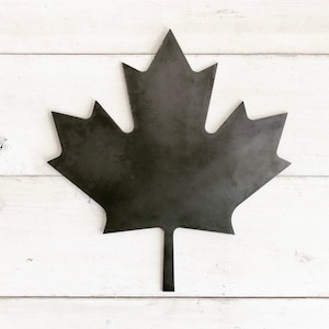 May include: A black metal maple leaf cutout, a popular symbol of Canada, is displayed against a white wood background.
