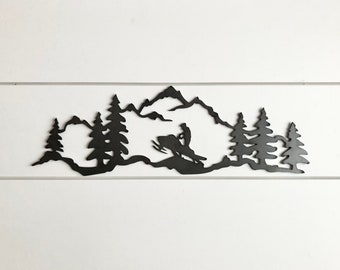 May include: Black metal wall art depicting a snowmobiler riding through a mountain landscape with pine trees.