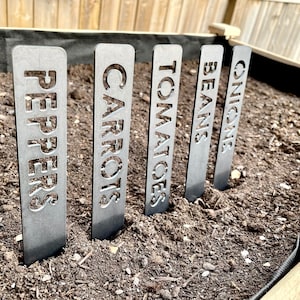 Peut inclure: Cinq marqueurs de jardin en métal avec les mots "ONIONS", "BEANS", "TOMATOES", "CARROTS" et "PEPPERS" découpés. Les marqueurs sont dans un lit de jardin avec de la terre brun foncé.