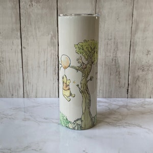 May include: A white tumbler featuring a Winnie the Pooh design. Pooh is holding a balloon and Piglet is in a tree. The tumbler has a green and brown background.