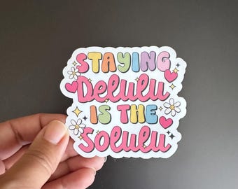 May include: A white sticker with the text "Staying Delulu is the Solution" in rainbow colours with hearts and flowers.