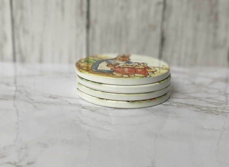 May include: Set of four white ceramic coasters featuring a vintage illustration of a rabbit family. The coasters are round and have a slightly raised edge.
