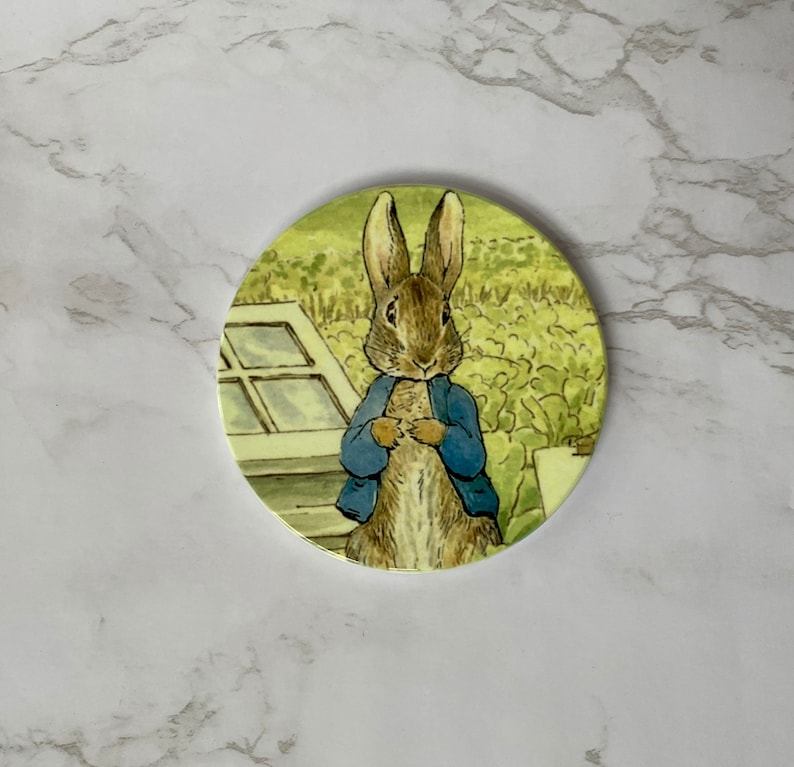 May include: A round coaster featuring a cartoon illustration of Peter Rabbit from Beatrix Potter's children's book. The coaster is white with a yellow background and depicts Peter Rabbit wearing a blue jacket and standing in a garden.