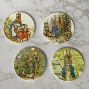 May include: Four round ceramic coasters featuring illustrations of Peter Rabbit and his family. The coasters are white with a green, brown, and blue colour scheme. The illustrations depict Peter Rabbit in various scenes, including gardening, playing with his siblings, and hiding in a garden.