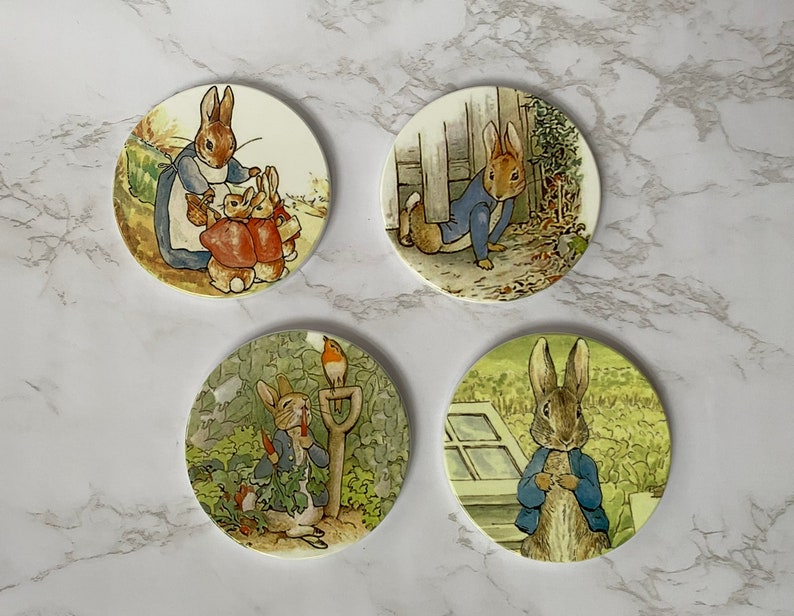 May include: Four round ceramic coasters featuring illustrations of Peter Rabbit and his family. The coasters are white with a green, brown, and blue colour scheme. The illustrations depict Peter Rabbit in various scenes, including gardening, playing with his siblings, and hiding in a garden.