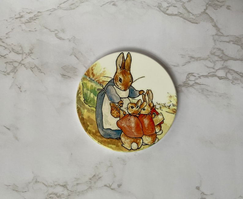 May include: A round ceramic coaster featuring a colourful illustration of a mother rabbit with two baby rabbits. The mother rabbit is wearing a blue dress and holding a basket. The baby rabbits are wearing red jackets. The coaster has a white background with a gold rim.