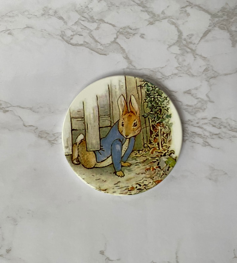 May include: A round ceramic coaster featuring a depiction of Peter Rabbit from Beatrix Potter's children's book. Peter is wearing a blue jacket and is peeking out from behind a white door.