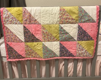 Op de afbeelding: Een roze en gele patchwork babydeken met een bloemenprint. De deken hangt aan een wieg met een roze ruche.