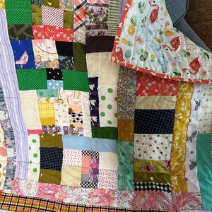 Può includere: Una trapunta patchwork con una varietà di motivi e colori di tessuto. La trapunta presenta quadrati, strisce e disegni floreali nei toni del verde, rosa, giallo e blu. Il bordo ha un motivo con fenicotteri e ananas.