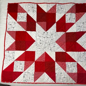 May include: A handmade quilt featuring a large star design. The quilt is primarily red and white, with various patterns including solid red, small red dots, and a white fabric with black scissors. The quilt has a red border.