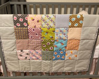 Op de afbeelding: Een patchwork babydeken met verschillende bloemenpatronen in tinten roze, groen, blauw en bruin. De deken hangt aan een wieg.
