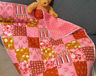 Op de afbeelding: Een roze en oranje patchwork babydeken met een teddybeer die eronder uitpiept.