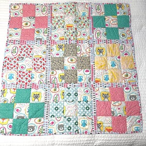 May include: A patchwork quilt with a variety of patterned squares in pink, teal, yellow, and cream. The squares feature floral, animal, and geometric designs. The quilt is bordered with a pink and white striped fabric.