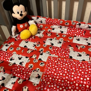May include: A red and white crib bedding set with a Minnie Mouse theme. The set includes a quilt with a patchwork design featuring Minnie Mouse faces and polka dots, and a separate pillow with a polka dot pattern.