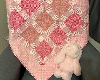 Op de afbeelding: Een roze en witte patchworkdeken met een roze pluche konijnenspeelgoed erop. De deken heeft een ruitpatroon en is gemaakt van verschillende stoffen met roze en witte bloemenmotieven.