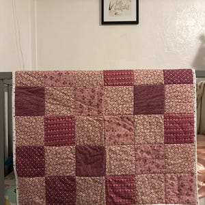Peut inclure: Une courtepointe patchwork rose et bordeaux, avec des carrés à motifs floraux. La courtepointe est posée sur un lit bébé gris. Une affiche encadrée avec l'inscription "hello beautiful" est accrochée au mur.