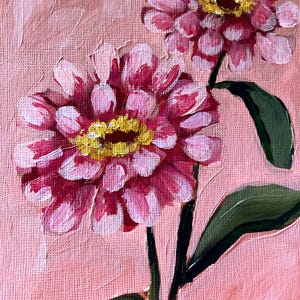 May include: An original oil painting of two pink zinnias with yellow centers and green leaves against a pink background. The flowers have multiple layers of petals, and the brushstrokes are visible, giving the painting a textured appearance.