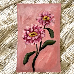 May include: An original painting of two pink zinnia flowers with yellow centers and green leaves on a pink background. The artwork is on a rectangular canvas. The painting is an oil painting.