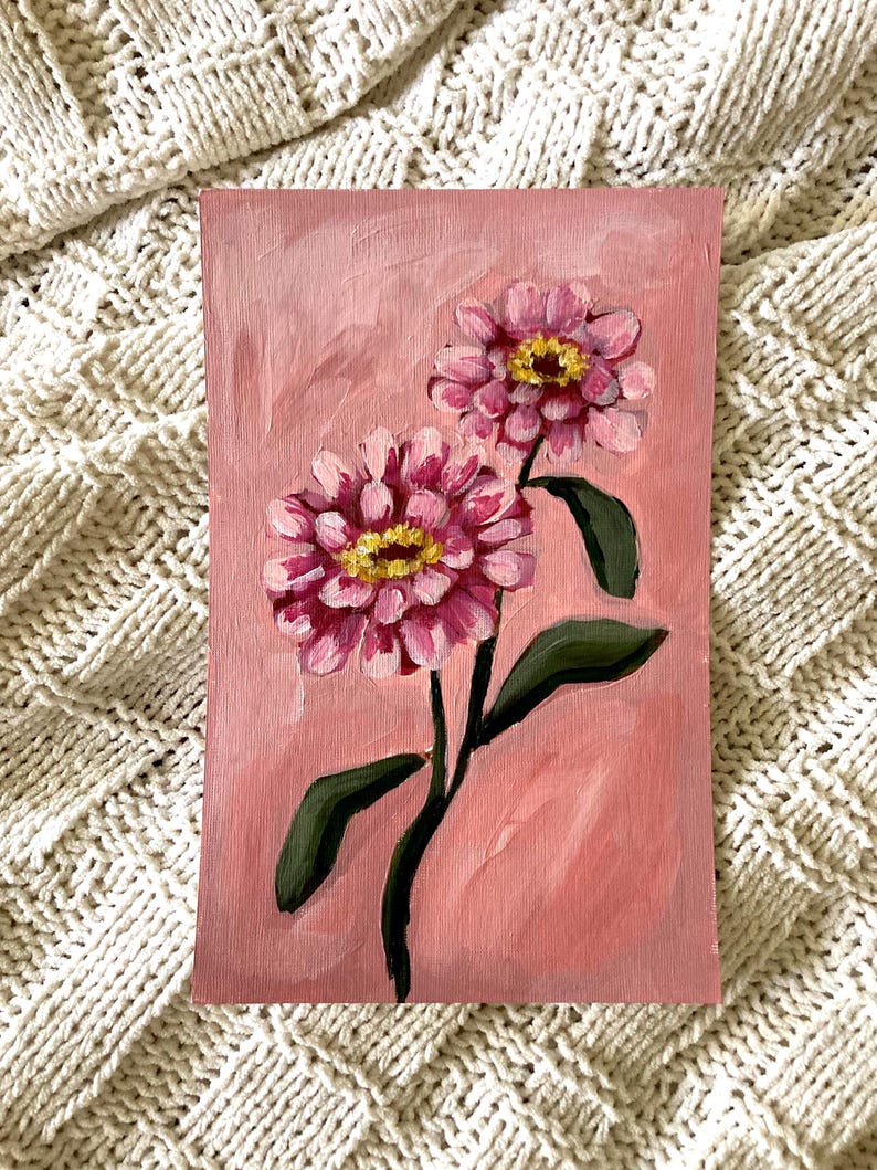 May include: An original painting of two pink zinnia flowers with yellow centers and green leaves on a pink background. The artwork is on a rectangular canvas. The painting is an oil painting.