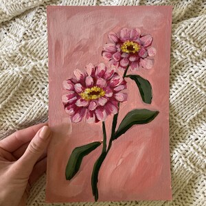 May include: A pink floral painting on canvas. Two pink and yellow zinnias are depicted with green leaves and stems against a pink background. The artwork is an original painting.