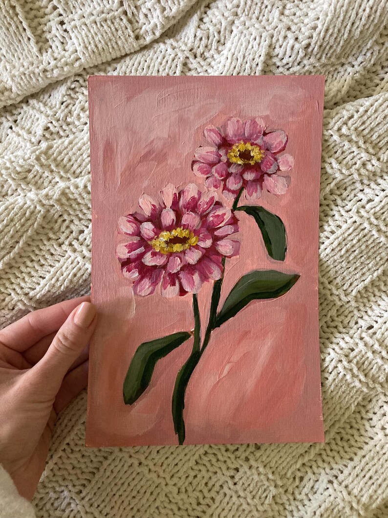 May include: A pink floral painting on canvas. Two pink and yellow zinnias are depicted with green leaves and stems against a pink background. The artwork is an original painting.