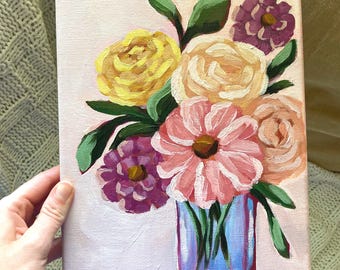 May include: An acrylic painting on a rectangular canvas featuring a bouquet of flowers in a blue glass vase. The arrangement includes yellow, peach, pink, and purple blossoms with green leaves, all set against a soft, light pink background.