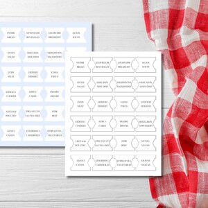 May include: Two sheets of pantry labels with various food categories printed in black. The labels are white with a light blue background. A red and white checkered cloth is in the background.