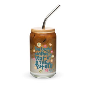 Puede incluir: Una lata de vidrio con tapa de bambú y una pajita. La lata está llena de café helado y tiene el texto "She laughs without fear of the future" y un versículo bíblico impreso.