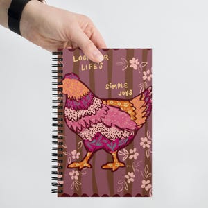 May include: A spiral bound notebook with a pink and orange patterned chicken illustration. The background is a burgundy color with brown stripes and pink flowers. The text on the notebook reads "Look for life's simple joys."