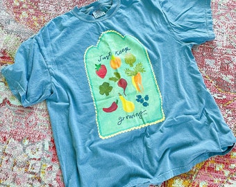 May include: A light blue t-shirt featuring a graphic of a green sign with the text "Just Keep Growing" and various colourful fruits and vegetables.