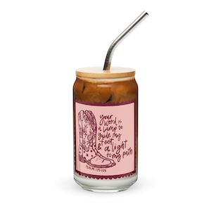 May include: A glass tumbler with a wooden lid and a stainless steel straw. The tumbler features a pink label with a drawing of a cowboy boot and the text "your word is a lamp to guide my feet & a light to my path".